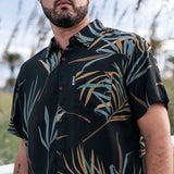 Man wearing a black short-sleeve flomotion button up with tropical leaf pattern outdoors.