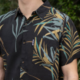Person wearing a black flomotion hirt with tropical leaf pattern in front of green foliage